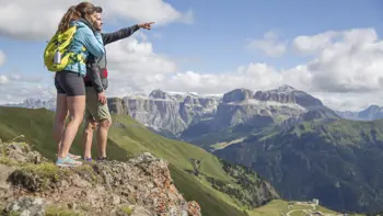People enjoy a breathtaking walk along the Belvedere viewpoint in Canazei, Val di Fassa, surrounded by the dramatic, jagged peaks of the Dolomites under a bright sky.