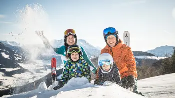 A family stand together in Kitzbuhel at the top of a mountain peak, playing with the snow as they enjoy the breath-taking views.