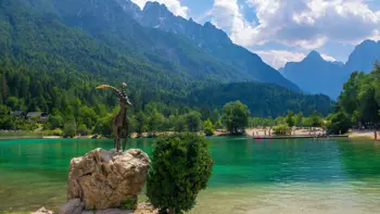 Steinbock (ibex) statue at Lake Jasna in Kranjska Gora, set on a stone platform beside the clear lake with alpine mountains and forests in the background.