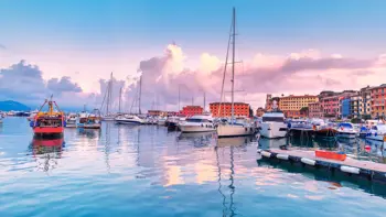 A collection of boats resting in the sunny waters of a Liguria harbour, with a collection of colourful buildings behind them.
