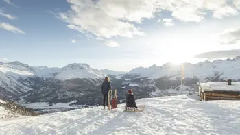 A couple stand atop a mountain in St. Moritz, ready with a pair of sleds to rush down the slopes ahead. They are surrounded by vast peaks and valleys.