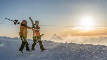 A couple of skiers walk along the top of a snowy peak, watching the sunset as they take in the beautiful views of mountains and valleys.