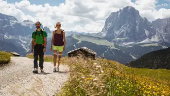 A couple walks  along a scenic trail in the Dolomite mountains near Selva, with towering limestone peaks and alpine meadows in the background.