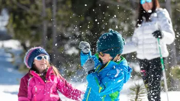 Two children play in the winter amongst themselves, kicking up powdered snow whilst surrounded by lush forests.