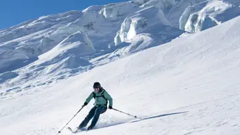 A lone skier kicks up powered snow as they rush down the side of a white mountain, surrounded by white peaks and valleys.